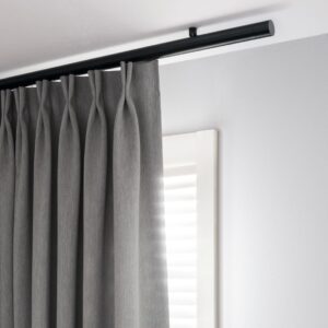 Moderne Interstil Vorhangstange „Corpus“ in mattem Schwarz, montiert direkt unter der Decke. Die runde, minimalistische Schiene trägt einen grauen Vorhang mit elegantem Faltenwurf (Flämische Falte). Das Design besticht durch eine klare, reduzierte Linienführung in einer hellen, zeitgemäßen Raumsituation.