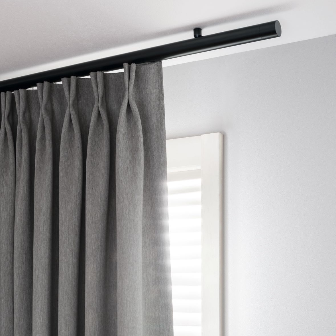 Moderne Interstil Vorhangstange „Corpus“ in mattem Schwarz, montiert direkt unter der Decke. Die runde, minimalistische Schiene trägt einen grauen Vorhang mit elegantem Faltenwurf (Flämische Falte). Das Design besticht durch eine klare, reduzierte Linienführung in einer hellen, zeitgemäßen Raumsituation.
