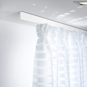 Puristisches Interstil Schienensystem „Helix“ in mattem Weiß, bündig an einer weißen Decke montiert. Die extrem flache, rechteckige Vorhangschiene trägt einen weißen, halbtransparenten Vorhang mit Querstreifen-Struktur, der in dekorativen Falten fällt. Das Bild betont eine helle, lichtdurchflutete Atmosphäre mit sanften Lichtreflexionen an der Decke.