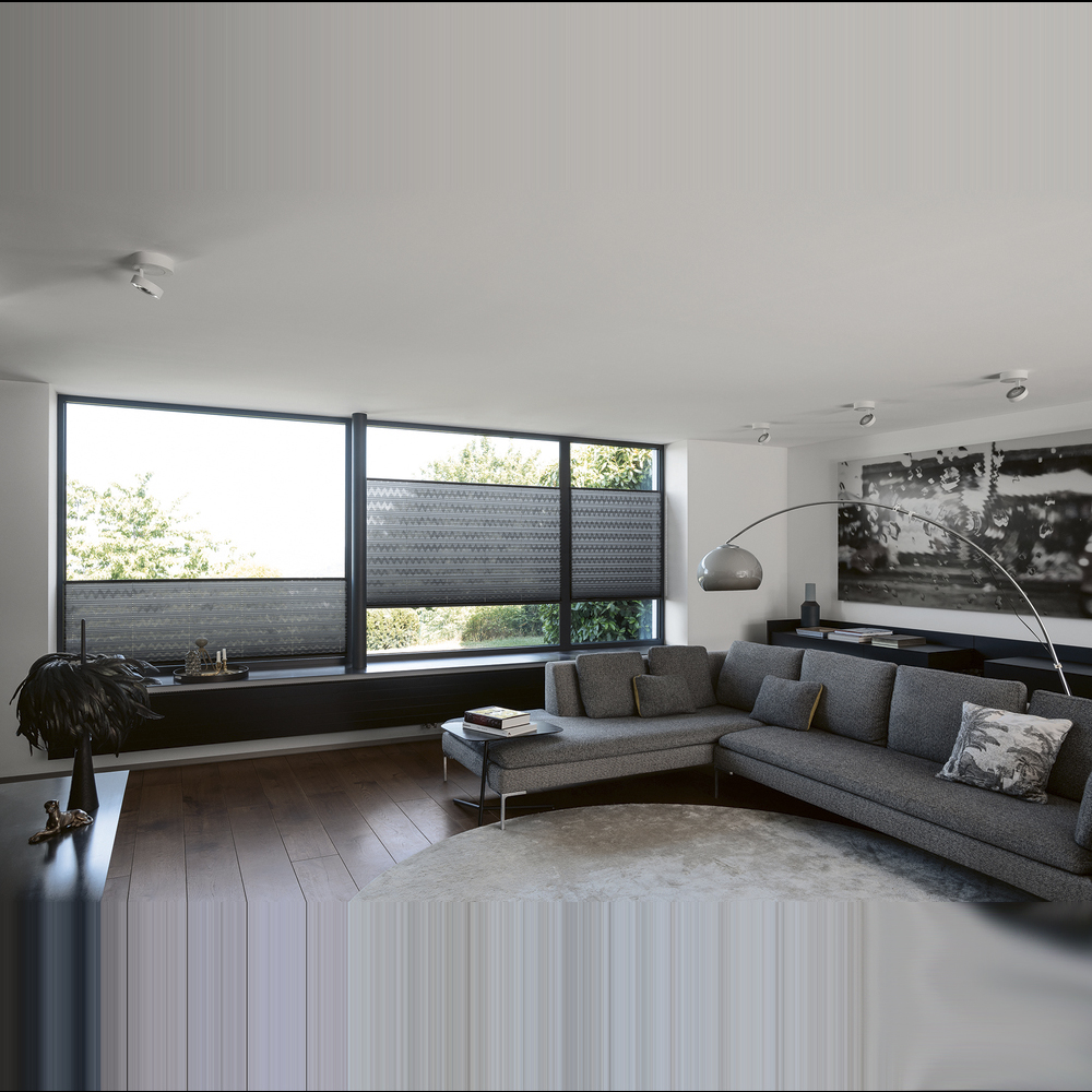 Blick durch ein großes Fenster mit einem grauen, horizontal gestreiften Plissee auf einen herbstlichen Wald. Das Plissee ist als Sichtschutz im unteren Fensterdrittel positioniert und erzeugt durch die Webstruktur ein elegantes Lichtspiel.