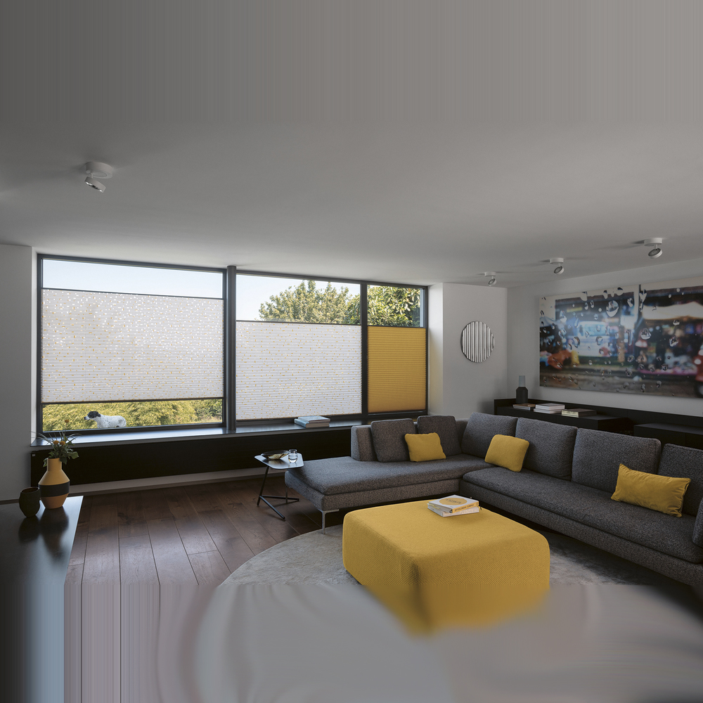 Nahaufnahme eines dunkelgrauen MHZ Plissees mit einer markanten, groben Webstruktur. Das Sonnenlicht fällt seitlich auf den Stoff und hebt die dreidimensionale Textur des Materials hervor, während im Hintergrund ein helles Fenster und eine Zimmerpflanze angedeutet sind.