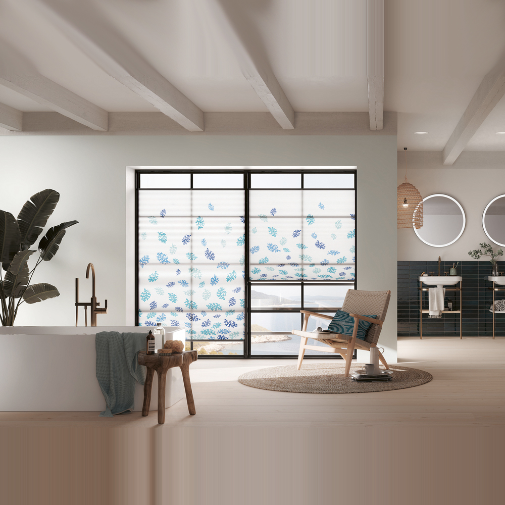 „Ein hochwertiges MHZ Wabenplissee aus der 'sieger design' Kollektion in einem Badezimmer. Das Plissee hat ein dezentes Wellenmuster in Blau-Grau (Design 'elements water') und ist im unteren Teil des Fensters als Sichtschutz positioniert, während oben der Blick ins Grüne frei bleibt.