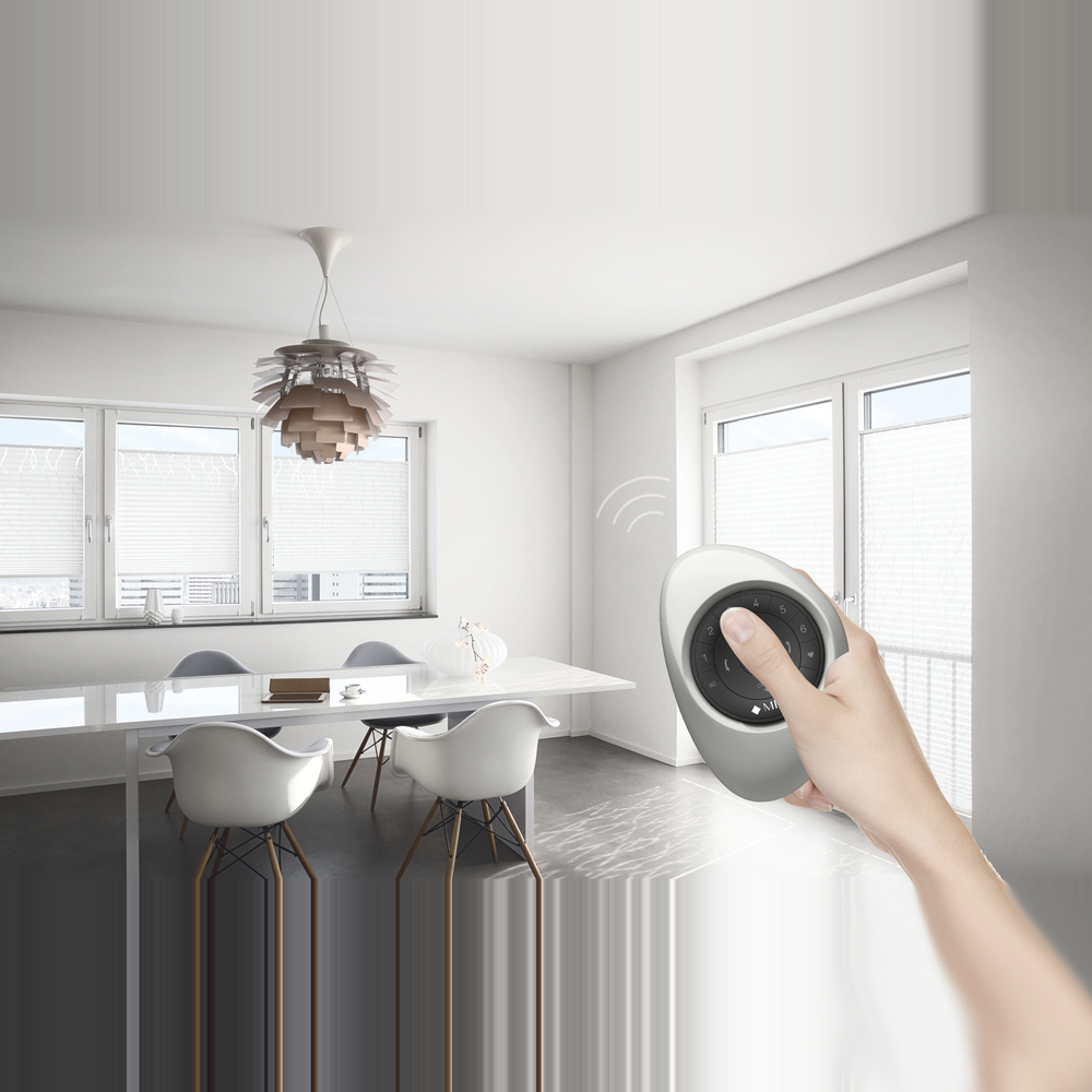 „Ein hellgraues MHZ Plissee an einem Fenster, das bequem per Fernbedienung gesteuert wird. Eine Hand hält die weiße PowerView-Fernbedienung ins Bild, während im Hintergrund ein minimalistisch eingerichteter Raum mit einer Pflanze zu sehen ist.