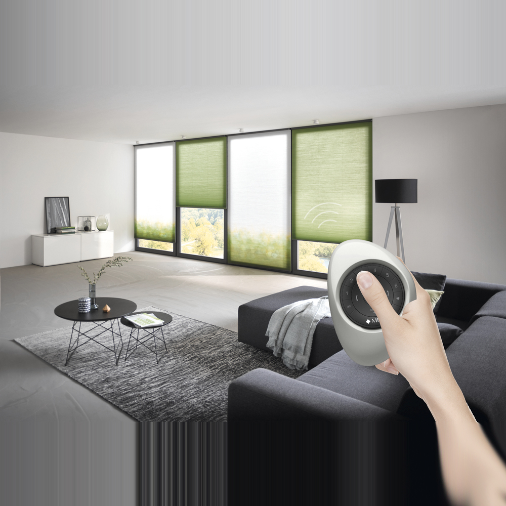 „Ein graues MHZ Wabenplissee an einem Fenster, das über die PowerView-Technologie gesteuert wird. Eine Hand hält die passende weiße Fernbedienung ins Bild. Das Waben-Muster des Stoffes ist in der Nahaufnahme gut erkennbar und filtert das Tageslicht sanft.