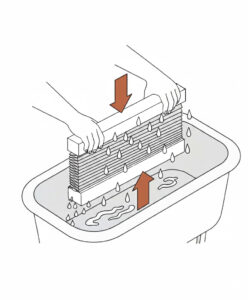 Eine schematische Illustration zeigt die Handwäsche eines kompakten Wabenplissees in einer Badewanne. Zwei Hände halten die Schienen des Plissees fest, während es senkrecht in das Wasserbad eingetaucht wird. Ein brauner Pfeil, der nach unten zeigt, und ein brauner Pfeil, der nach oben zeigt, verdeutlichen das vorsichtige Schwenken des Stoffes im Wasser, um Verschmutzungen zu lösen.