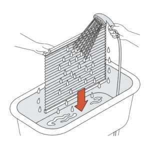 Eine schematische Strichzeichnung zeigt, wie ein Plissee in einer Badewanne gereinigt wird. Eine Person hält das auseinandergezogene Plissee senkrecht über die Wanne, während eine zweite Hand es mit einem Duschkopf gründlich abspült. Ein großer roter Pfeil zeigt nach unten in die Badewanne, um das Ablaufen des Wassers zu verdeutlichen.