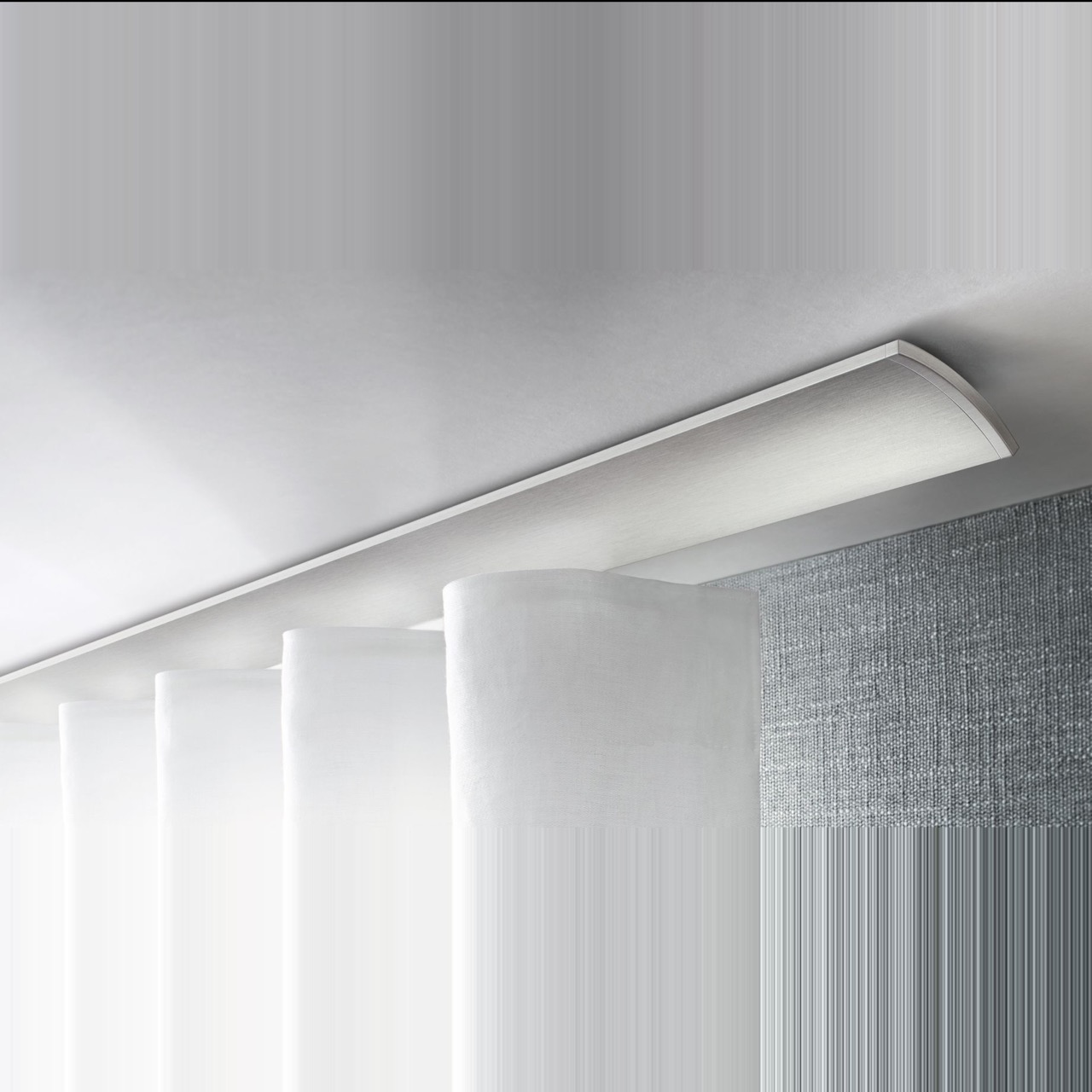 Architektonisches Interstil Schienensystem „W2“ in edlem, matt gebürstetem Aluminium. Die flache Schiene weist eine sanft nach außen gewölbte (konvexe) Front auf und ist direkt an der Decke montiert. Sie trägt einen weißen, blickdichten Wellenvorhang, der fließend unter dem Profil hervortritt. Das Bild vermittelt eine hochwertige, moderne Ästhetik vor einer strukturierten, grauen Wandfläche.